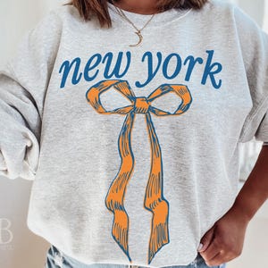 May include: A gray sweatshirt with the text "new york" in blue lettering and a large orange bow with blue outlines.