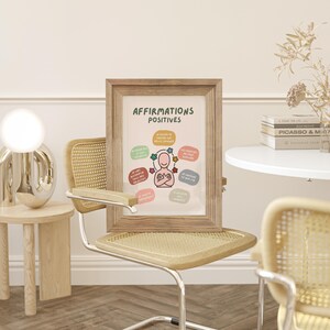 May include: A framed print with the words "AFFIRMATIONS POSITIVES" in a wooden frame. The print features a cartoon figure surrounded by colorful affirmations. The print is displayed on a light-colored chair.