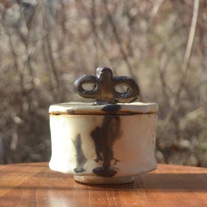 Puede incluir: Un pequeño tarro de cerámica hecho a mano con tapa. El tarro es de color blanquecino con gotas de color marrón oscuro y un asa decorativa en la parte superior. Se encuentra sobre una mesa de madera, con un fondo natural borroso.