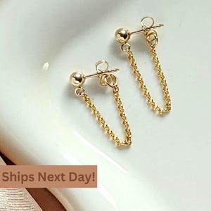 May include: A pair of gold chain earrings with small round balls at the top. The chains are connected to the balls and hang down.
