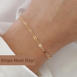 May include: A gold chain bracelet with a delicate, rectangular link design. The bracelet is shown on a person's wrist.