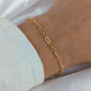 May include: A gold-toned bracelet featuring a delicate paperclip chain design. The chain is composed of elongated, rectangular links connected by smaller circular links, creating a refined and uniform appearance that rests against the wrist.