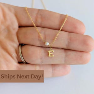 May include: A delicate gold-colored necklace featuring a small letter "E" pendant and a single pearl. The necklace is displayed on a hand, showcasing its fine chain and minimalist design. The jewelry is a personalized accessory.