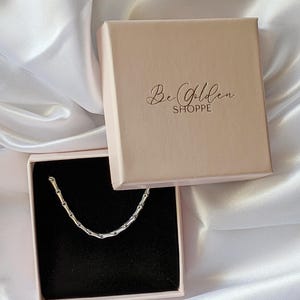 Puede incluir: Un collar de cadena plateado en una caja rosa con el texto "Be Golden Shoppe" en la tapa.