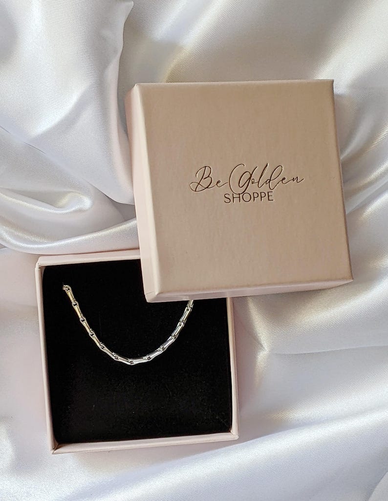 Puede incluir: Un collar de cadena plateado en una caja rosa con el texto "Be Golden Shoppe" en la tapa.