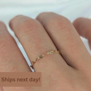 May include: A thin gold ring featuring a delicate chain-like design composed of small, circular, textured links with a sunburst pattern. The ring is worn on a finger, showcasing its fine, metallic construction and intricate, repeating circular details.
