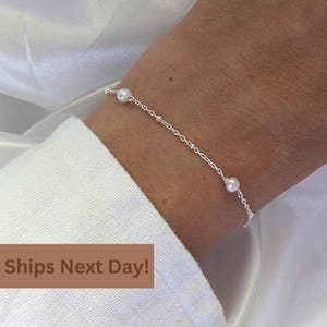 May include: A delicate silver bracelet with three white pearls and small silver beads. The bracelet is worn on a wrist, against a white fabric background. The bracelet is a simple, elegant piece of jewelry.