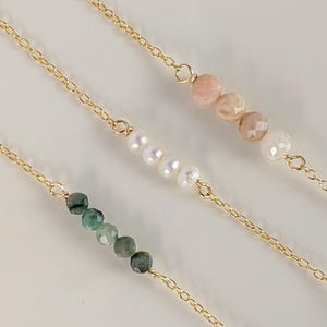May include: Three gold chains with different gemstone beads. The top chain has pink beads, the middle chain has white beads, and the bottom chain has green beads.