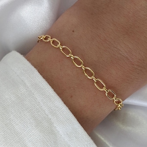 May include: A gold-colored bracelet with an oval link chain design. The bracelet is worn on a wrist, with a white shirt visible in the background. The bracelet is a simple, elegant piece of jewelry.