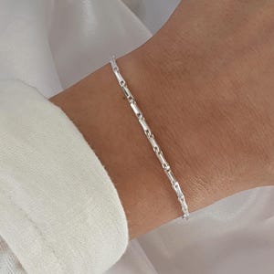 Puede incluir: Un brazalete de cadena de plata con un delicado dise&ntilde;o de eslabones cuadrados. El brazalete se muestra en la mu&ntilde;eca de una persona.
