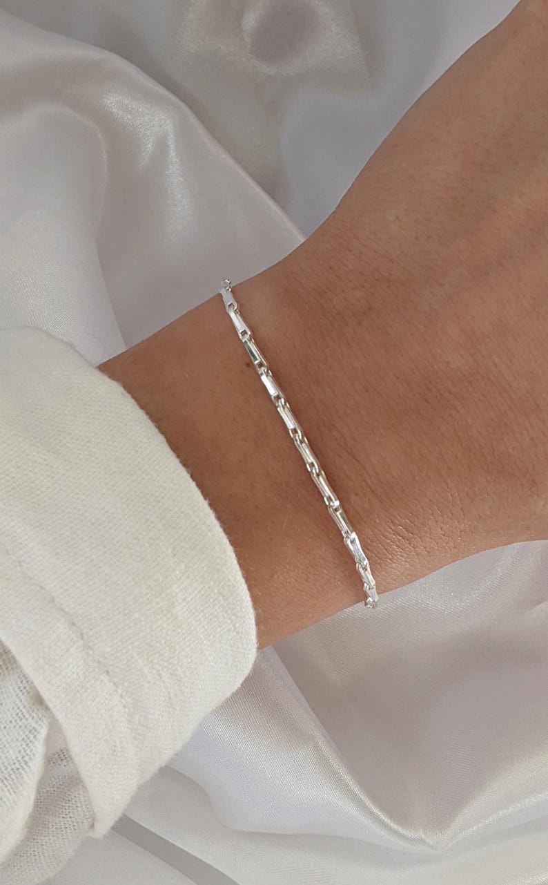 Puede incluir: Un brazalete de cadena de plata con un delicado dise&ntilde;o de eslabones cuadrados. El brazalete se muestra en la mu&ntilde;eca de una persona.