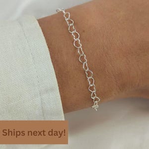 May include: A silver-toned chain bracelet composed of a series of interconnected heart-shaped links. The bracelet is worn around the wrist, showcasing a delicate and continuous pattern of small, uniform metallic hearts linked together in a row.