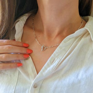 May include: A silver-colored necklace with a toggle clasp closure. The necklace is composed of small, rectangular links. The clasp is a circular bar that fits through a loop. The necklace is worn against a white shirt.