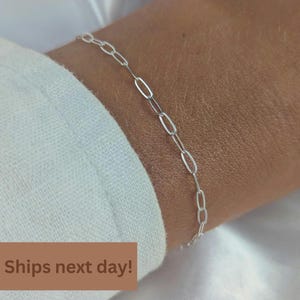 May include: A delicate silver chain bracelet featuring elongated, oval-shaped paperclip links. The bracelet is composed of polished metallic silver material, showcasing a minimalist design with a repeating pattern of open, rectangular links.