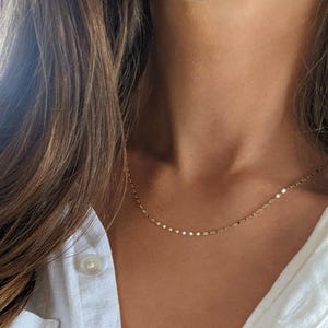 May include: Delicate gold necklace with small, round charms. The necklace is worn against a white button-down shirt. The jewelry is a simple, elegant accessory.