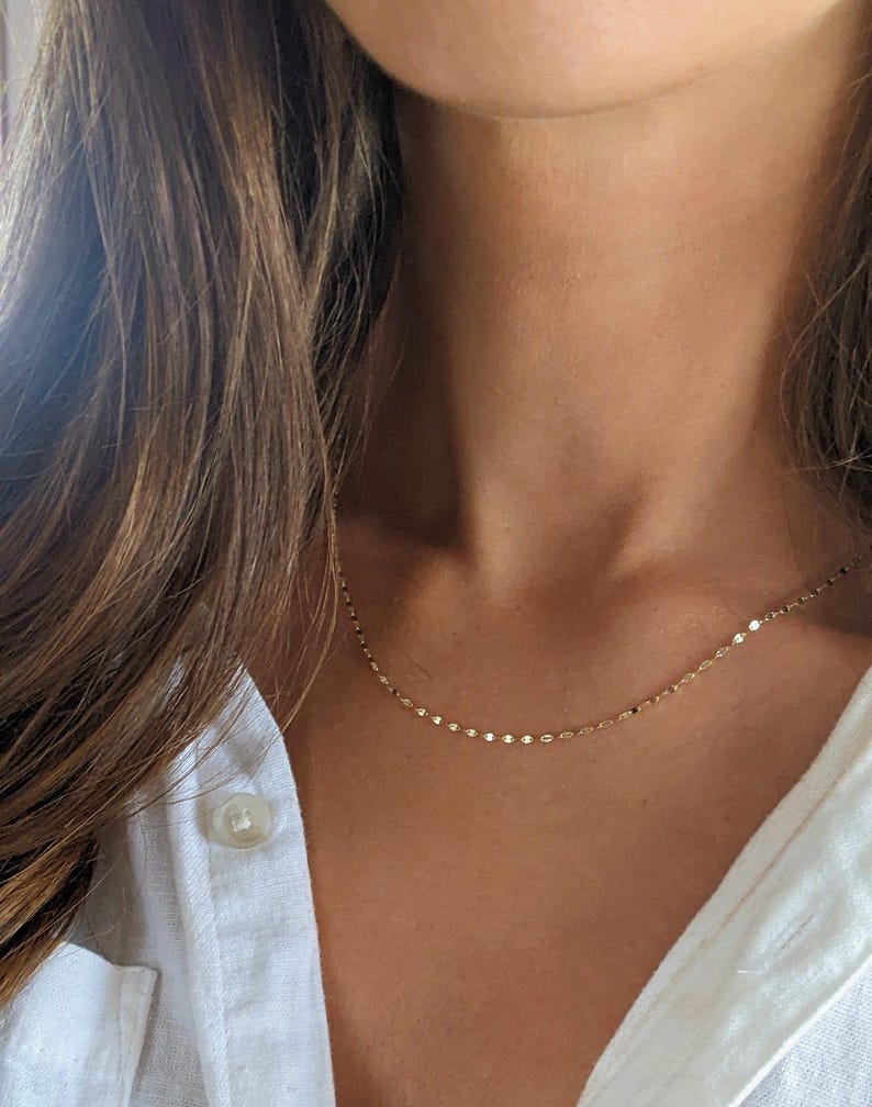 May include: Delicate gold necklace with small, round charms. The necklace is worn against a white button-down shirt. The jewelry is a simple, elegant accessory.