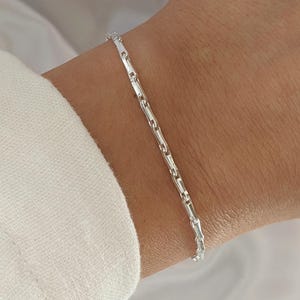 Puede incluir: Un brazalete de cadena de plata con un delicado dise&ntilde;o de cadena de eslabones cuadrados. El brazalete se muestra en la mu&ntilde;eca de una persona.