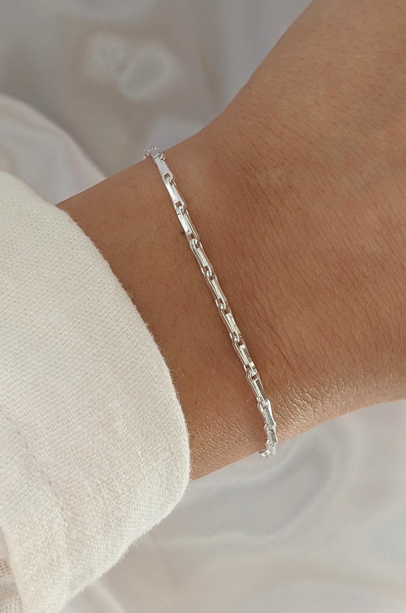 Puede incluir: Un brazalete de cadena de plata con un delicado dise&ntilde;o de cadena de eslabones cuadrados. El brazalete se muestra en la mu&ntilde;eca de una persona.