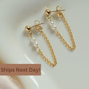May include: A pair of gold chain earrings with small white pearls hanging from the chains. The earrings are on a white surface.