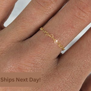 May include: A delicate gold ring featuring a continuous chain of small, interconnected heart-shaped links. The ring has a polished metallic finish and a thin, lightweight band design that encircles the finger, showcasing a repeating heart pattern.