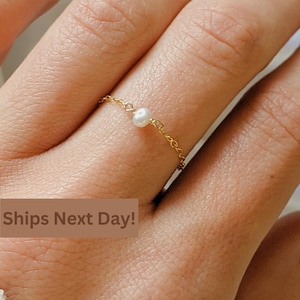 May include: A delicate gold chain ring with a single white pearl. The ring is worn on a finger.