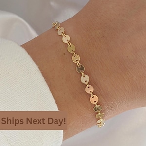 May include: A gold chain bracelet with small, round, flat discs. The bracelet is on a person's wrist.