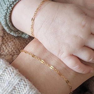 May include: Two gold chain bracelets with oval links. The bracelets are on the wrists of a person wearing a knitted sweater.