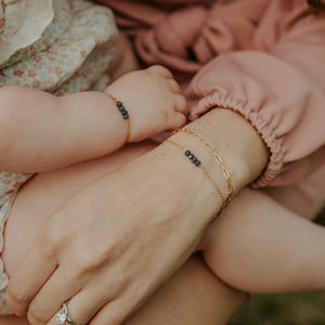 May include: A gold chain bracelet featuring four small dark beads on a wrist, paired with a second gold chain bracelet with elongated oval links. A matching gold chain bracelet with four dark beads is also visible on a smaller wrist nearby.