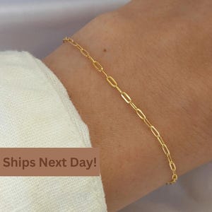 May include: A delicate gold-colored bracelet with an oval link chain. The bracelet is worn on a wrist, with a white shirt visible in the background. The text "Ships Next Day!" is displayed on a brown banner.