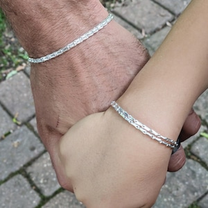 May include: Two silver bracelets, each with a linear design, are worn on wrists. The bracelets are made of small, rectangular, clear stones. The image shows two people holding hands, with the bracelets visible on their wrists.