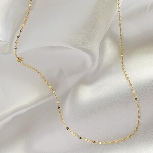 May include: Delicate gold necklace with a repeating oval link design. The necklace is displayed on a white, silky fabric, creating a contrast that highlights the jewelry's shine. A simple and elegant piece.