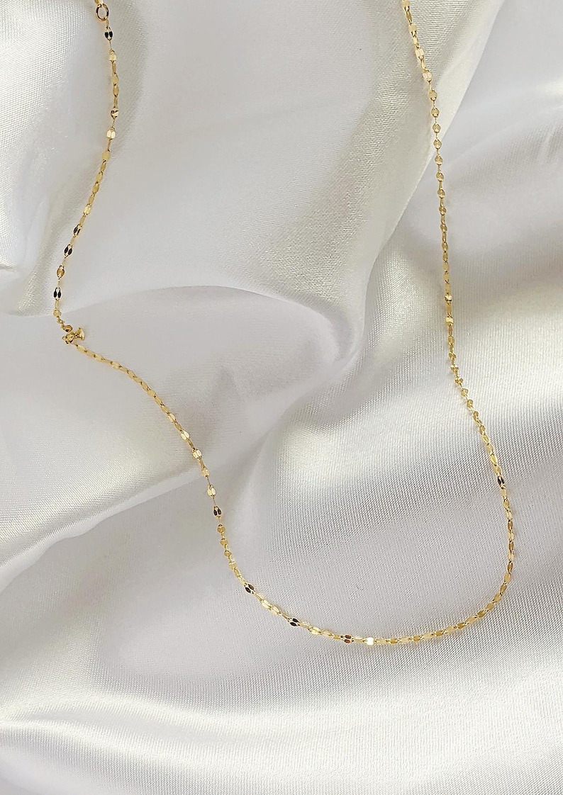 May include: Delicate gold necklace with a repeating oval link design. The necklace is displayed on a white, silky fabric, creating a contrast that highlights the jewelry's shine. A simple and elegant piece.