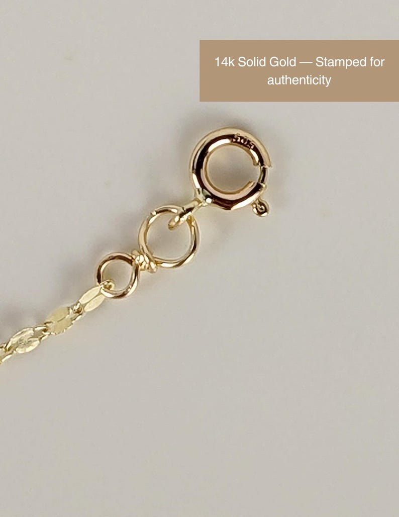 May include: Close-up of a 14k solid gold necklace clasp and chain. The clasp is round with a small lever, and the chain has a delicate, flat link design. Text reads: "14k Solid Gold &mdash; Stamped for authenticity."