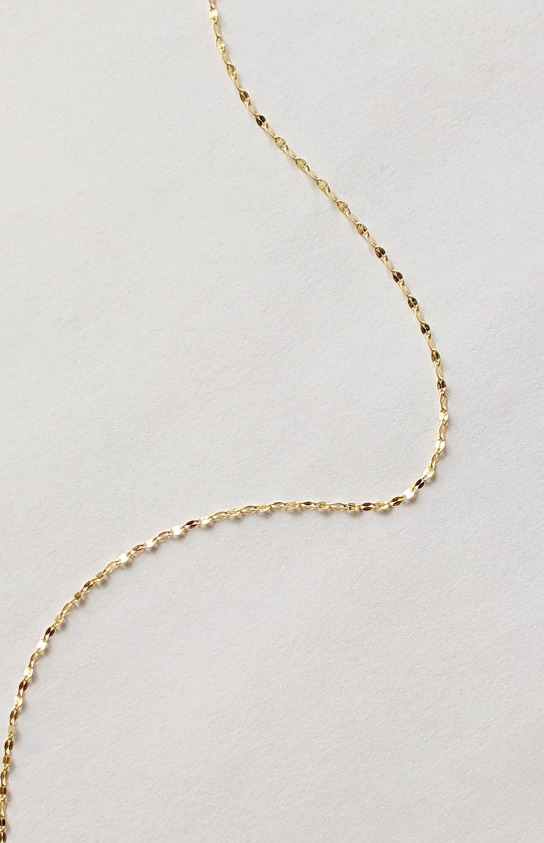 May include: A delicate gold-colored necklace with a repeating oval link chain design. The chain is displayed against a white background, highlighting its subtle shine and intricate pattern. This piece is suitable for everyday wear.
