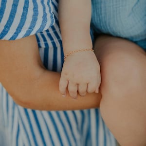 May include: A delicate gold-toned chain bracelet featuring a series of small, interconnected heart-shaped links. The bracelet is worn around a small wrist, showcasing the fine metal construction and the repeating pattern of the heart links.