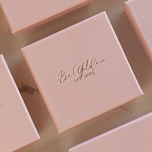 Puede incluir: Varias cajas cuadradas rosas con el texto "Be Golden Shoppe" en letras doradas.