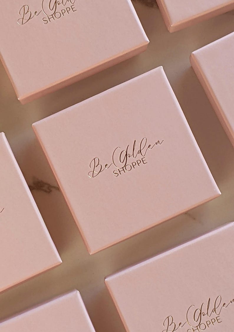 Puede incluir: Varias cajas cuadradas rosas con el texto "Be Golden Shoppe" en letras doradas.