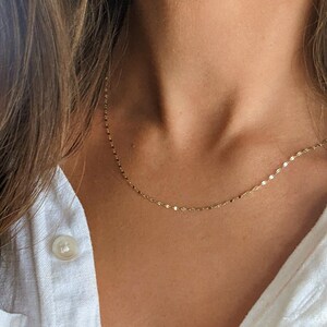 May include: Delicate gold necklace with a subtle, shimmering design. The necklace is shown against a person's skin and a white button-down shirt. The chain is made of small, flat, circular links.