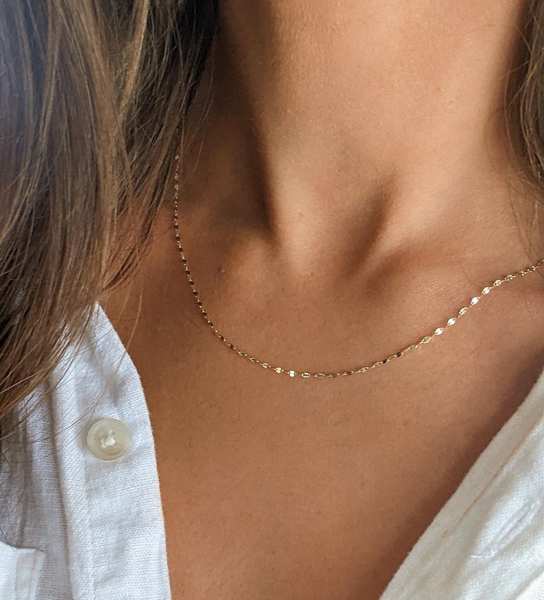 May include: Delicate gold necklace with a subtle, shimmering design. The necklace is shown against a person's skin and a white button-down shirt. The chain is made of small, flat, circular links.