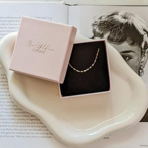 May include: A delicate gold necklace with a unique chain design rests inside a pink box. The box is open, revealing the necklace against a black interior. The box is on a white, curved tray, with text that reads "Be Golden SHOPPE".