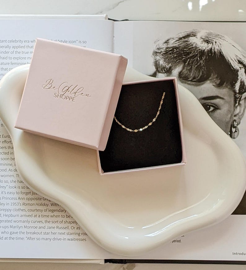 May include: A delicate gold necklace with a unique chain design rests inside a pink box. The box is open, revealing the necklace against a black interior. The box is on a white, curved tray, with text that reads "Be Golden SHOPPE".