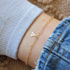 May include: A gold-colored anklet with a delicate heart chain design. The anklet features a small charm with the letter "A". The anklet is worn on an ankle with a denim jean and gray sock in the background.