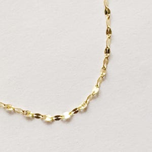 May include: Delicate gold necklace with a repeating, flattened oval link design. The chain is displayed against a white background, highlighting its subtle shine and intricate pattern. A simple, elegant piece of jewelry.