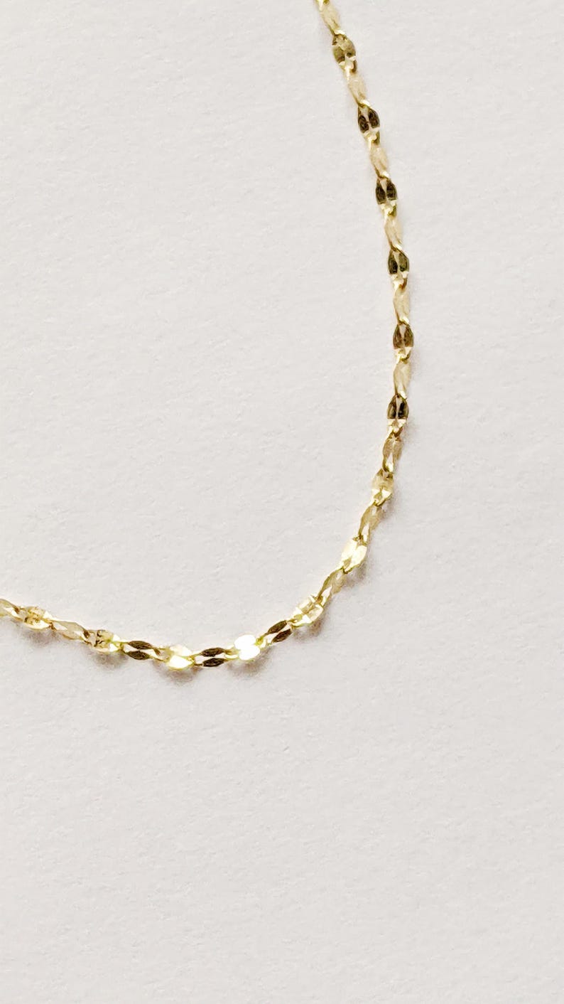 May include: Delicate gold necklace with a repeating, flattened oval link design. The chain is displayed against a white background, highlighting its subtle shine and intricate pattern. A simple, elegant piece of jewelry.