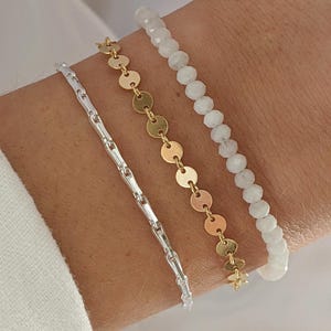 Puede incluir: Tres pulseras apiladas en una mu&ntilde;eca. La pulsera superior est&aacute; hecha de cuentas blancas. La pulsera del medio est&aacute; hecha de cuentas doradas en forma de monedas. La pulsera inferior es una cadena de plata.