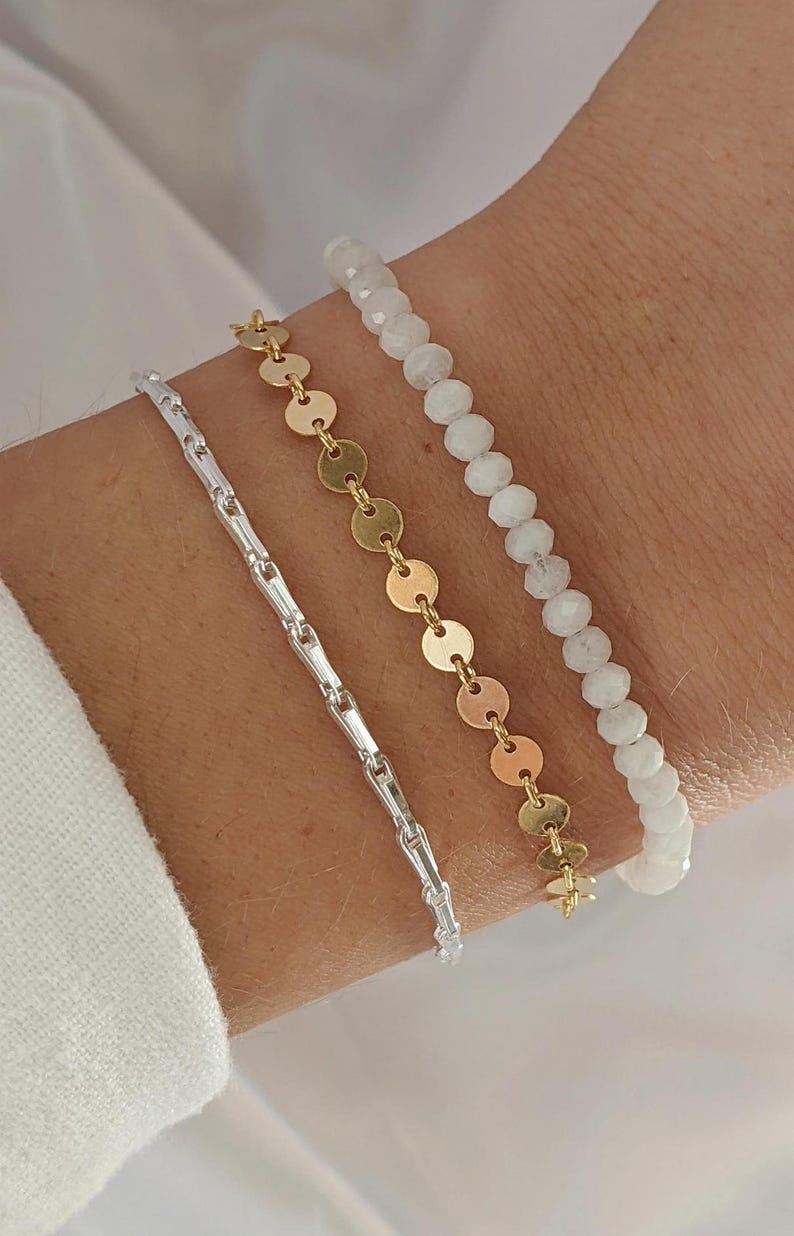Puede incluir: Tres pulseras apiladas en una mu&ntilde;eca. La pulsera superior est&aacute; hecha de cuentas blancas. La pulsera del medio est&aacute; hecha de cuentas doradas en forma de monedas. La pulsera inferior es una cadena de plata.