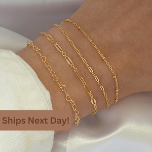May include: Four gold-colored bracelets with different chain designs are displayed on a wrist. The bracelets include heart-shaped links, oval links, rectangular links, and a beaded chain. The background is a white fabric.
