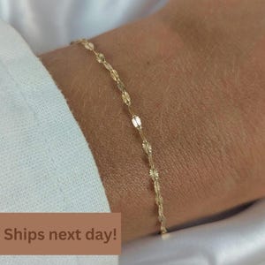 May include: A delicate gold chain bracelet featuring a series of small, flat, circular links with a textured, sunburst-like pattern. The bracelet is thin and minimalist in design, resting against the skin with a subtle, reflective metallic finish.