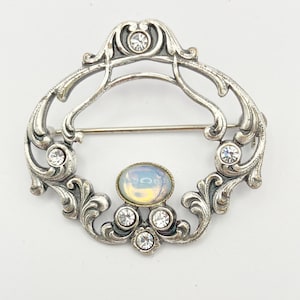 Pode incluir: Um broche oval de tom prateado com uma moldura de volutas intrincadas. A peça é decorada com seis pequenos strass redondos transparentes e uma pedra central oval em cabochão que exibe um brilho iridescente, branco leitoso e azul.