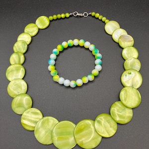 May include: A necklace composed of graduated, circular, lime green shell discs and a matching bracelet featuring small, round, multi-coloured beads in shades of white, yellow and green. The necklace includes a silver-toned metal lobster clasp.